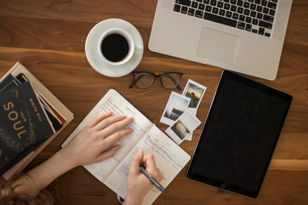 Table with items including books, photos, a cup of coffee, an ipad, a computer, and hands writing in a journal